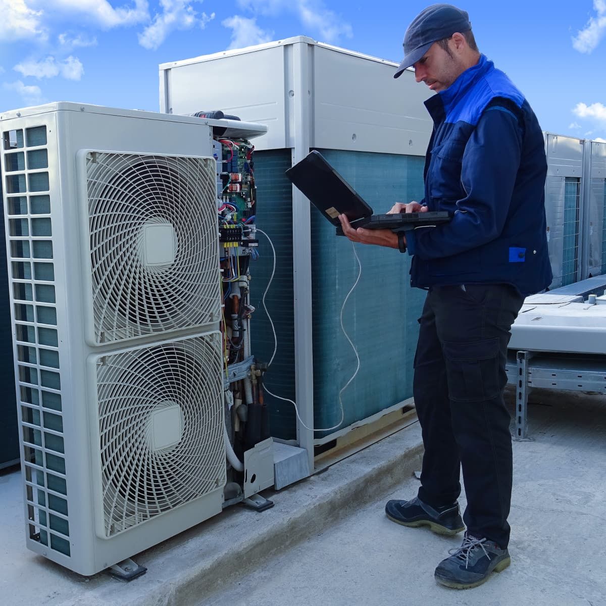 HVAC technician on a rooftop, using a laptop to inspect an air conditioning unit.