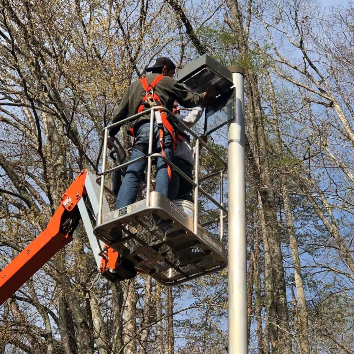 Two people in a lift bucket are working on a street light. Trees are in the background.