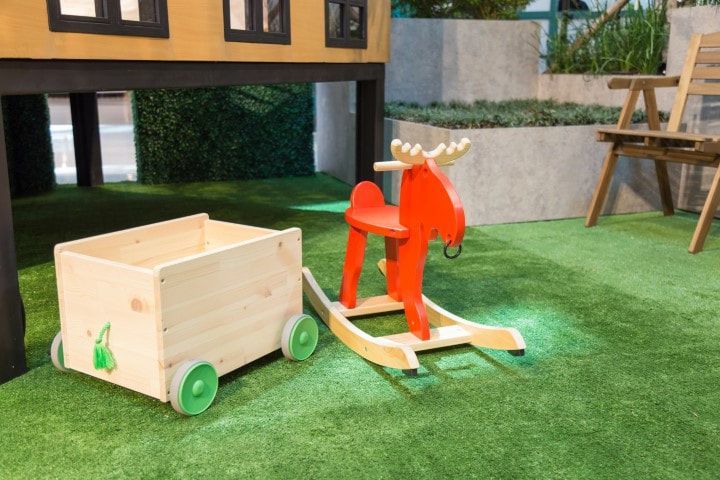Wooden toy cart and red rocking moose on green artificial grass near a building.