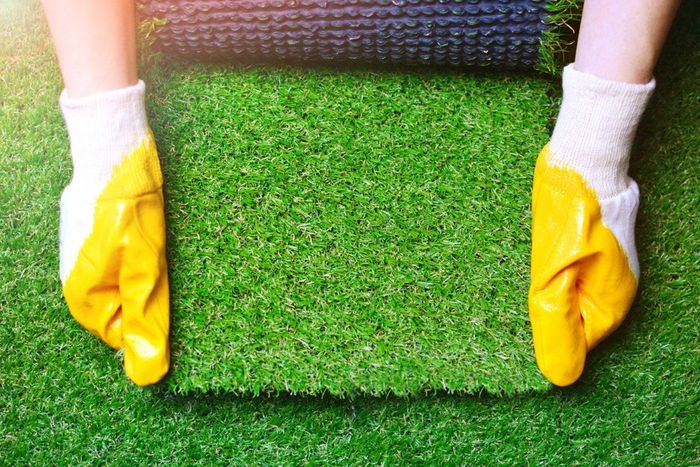 Hands in yellow gloves unroll a section of artificial turf onto a green surface.
