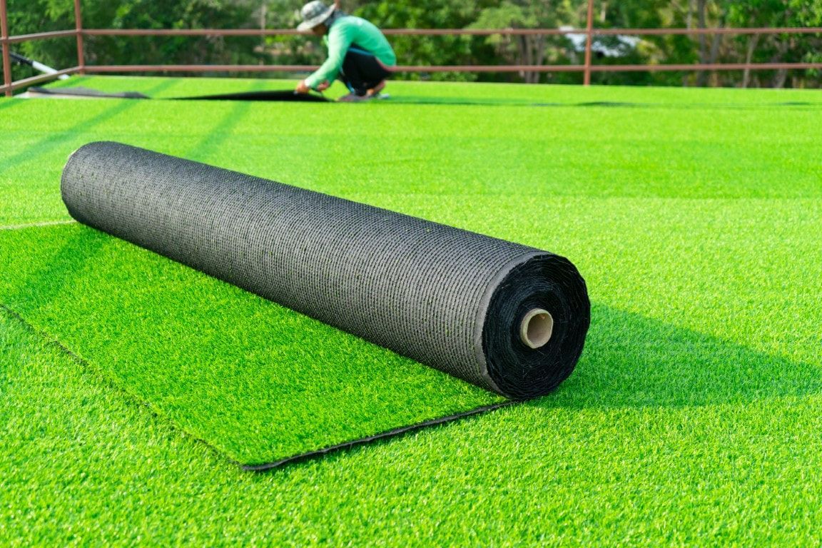 A roll of black underlayment and a section of artificial turf being installed on a sports field. A worker is in the background.