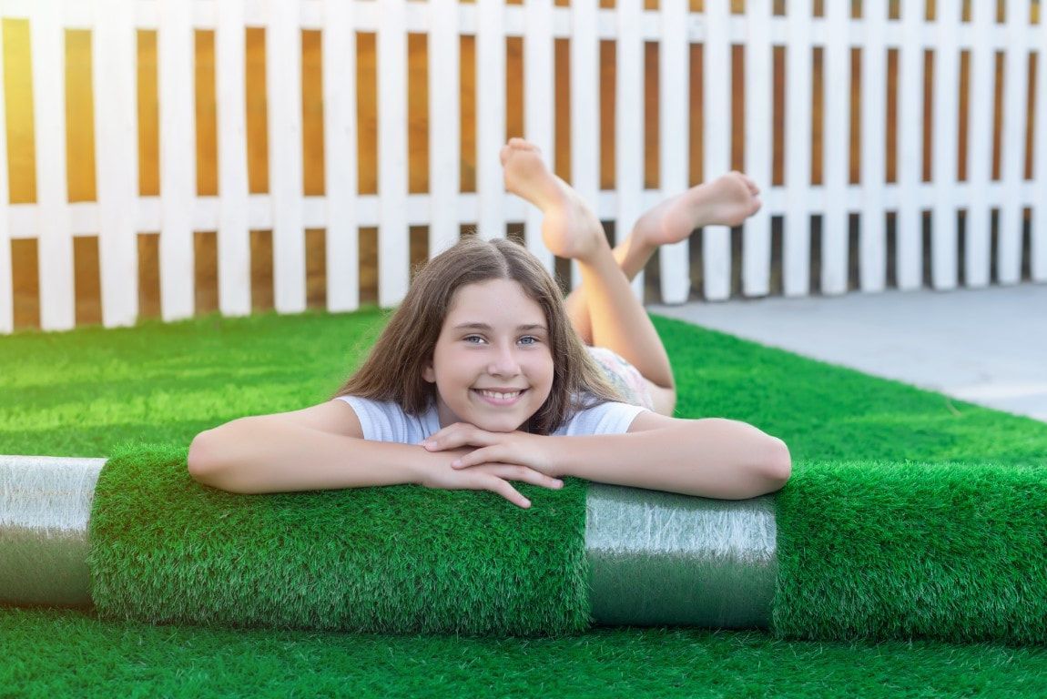 Girl smiles, lying on green artificial turf, arms crossed, feet up, white fence in background.