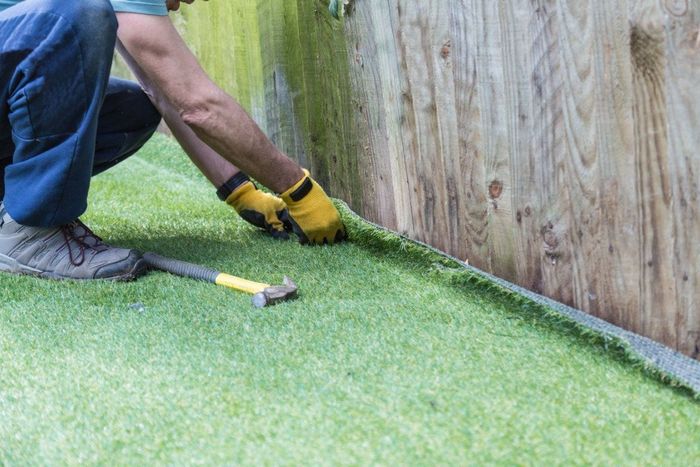 A person installing artificial turf near a wooden fence, wearing gloves and using a hammer.