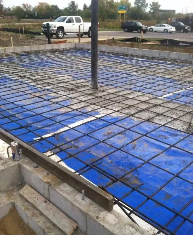 Construction site with rebar grid on blue tarp; concrete being poured from a pipe.