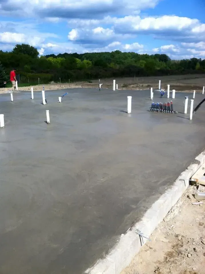Freshly poured concrete foundation with plumbing pipes, person in red shirt in background.