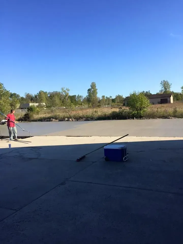 Man smoothing wet concrete with a long tool on a sunny day; cooler and tools in foreground.