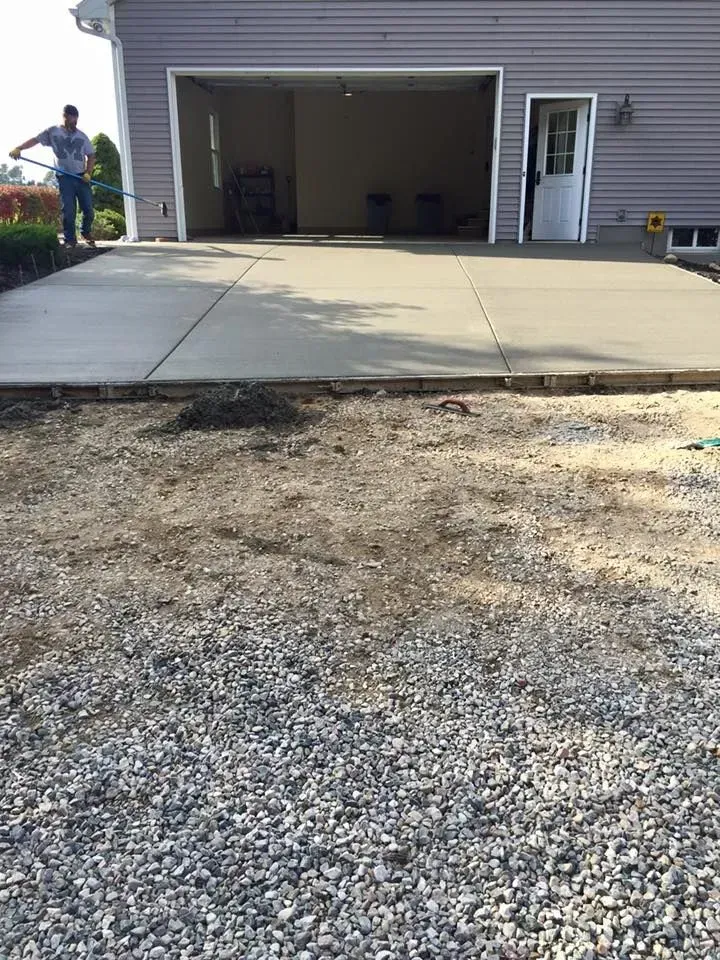 Man spraying concrete driveway in front of a garage with open doors and gravel area in the foreground.