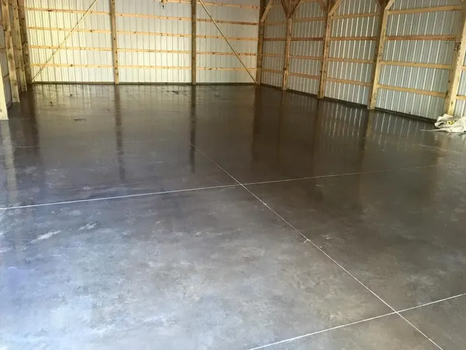 Interior of a large, empty concrete floored building with wooden framework and metal walls, reflecting light.