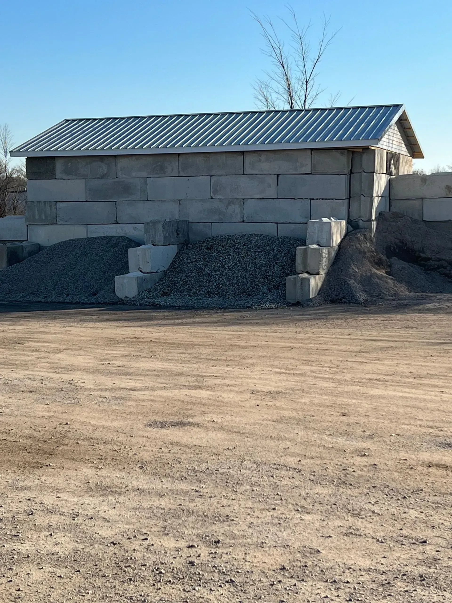 Gray concrete block building with a metal roof, next to a gravel pile, set on dirt ground.