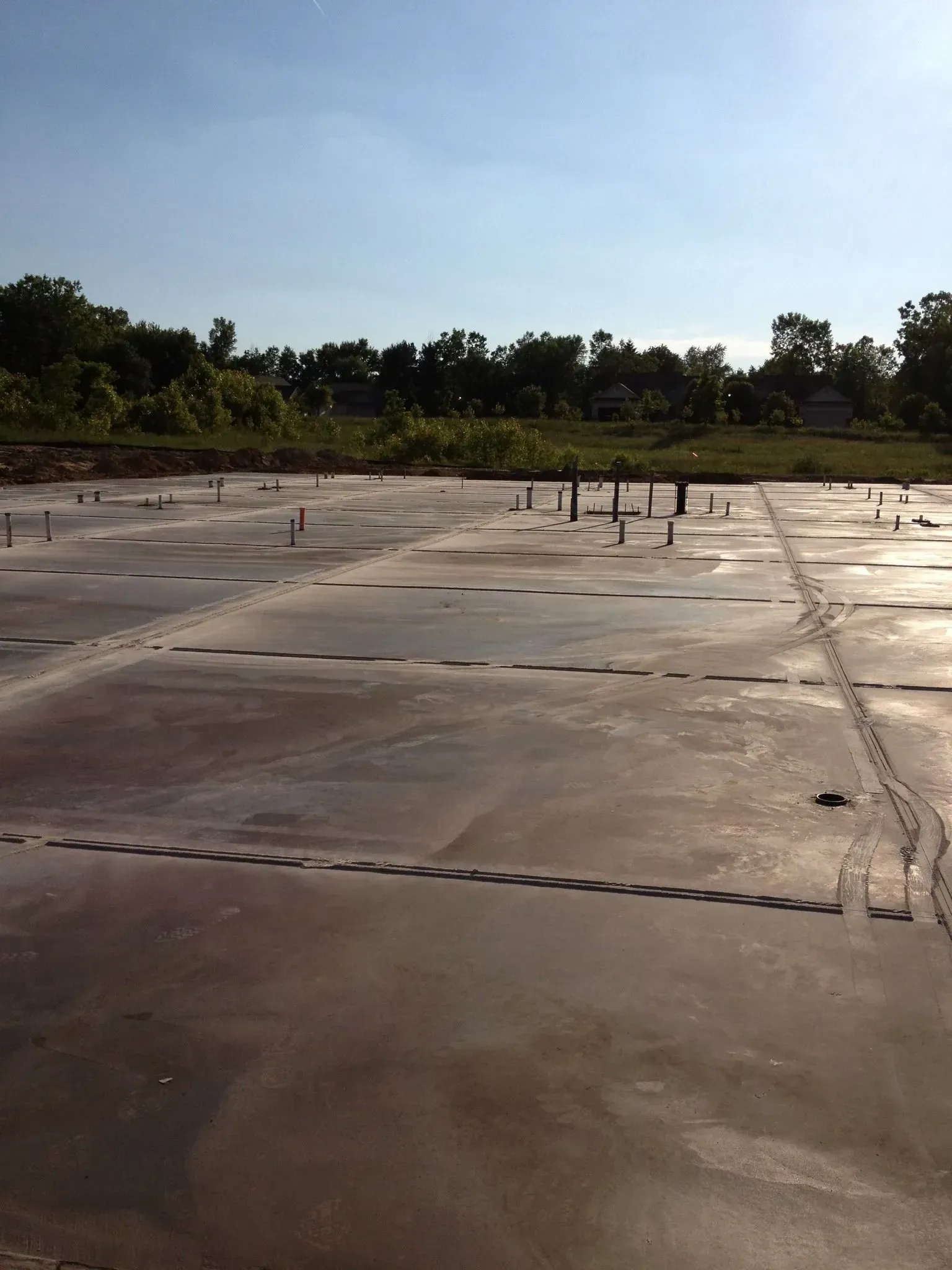 Newly poured concrete slab with embedded pipes and footprints, under a blue sky.