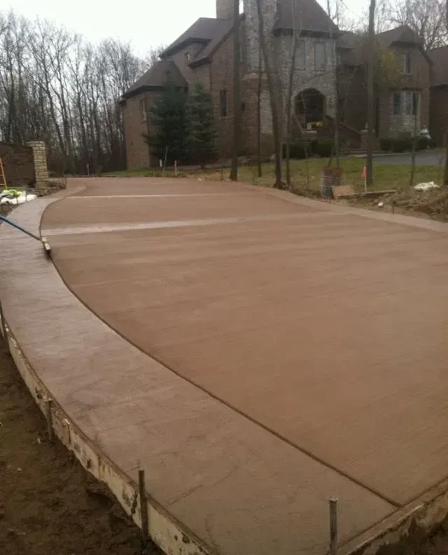 Newly poured concrete driveway with a large house in the background. Brown, flat surface with curved edges.