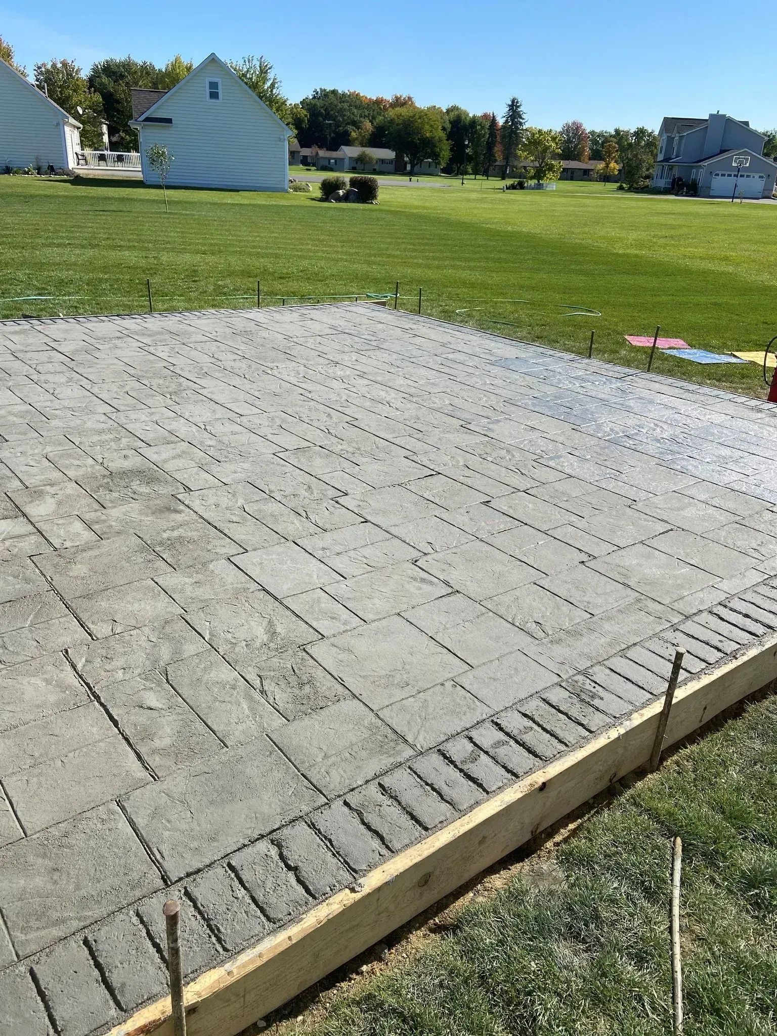 Freshly poured concrete patio with brick-like pattern, next to green grass and houses under a clear blue sky.
