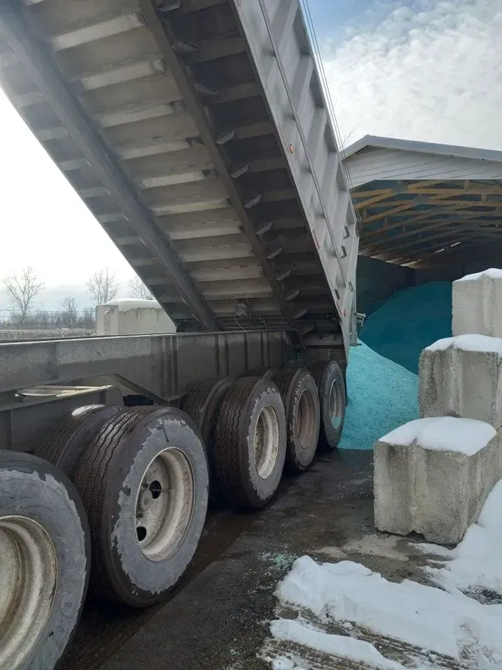 A dump truck unloading material into a blue pile, tires visible. Outdoors in a winter setting.