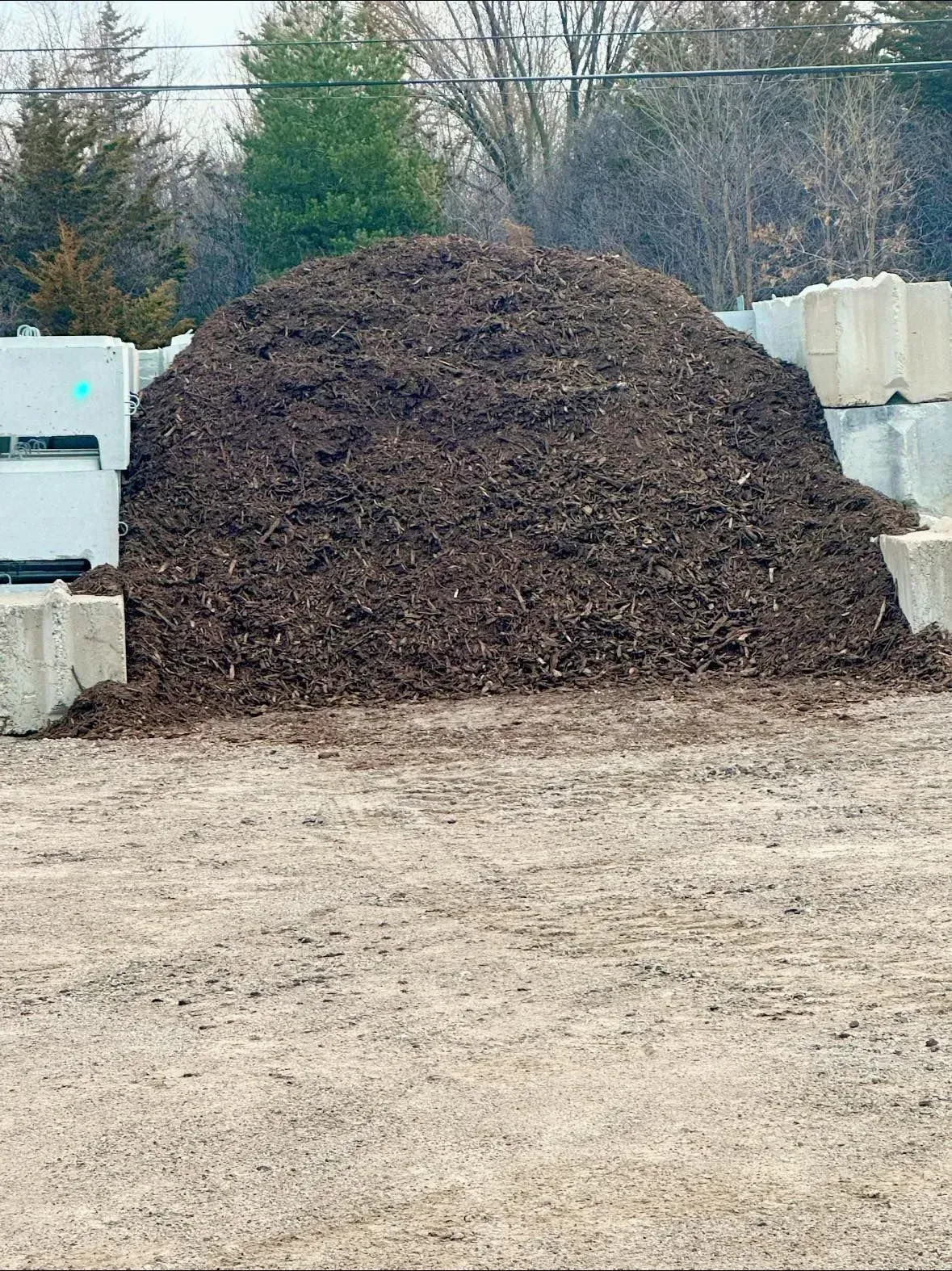 Pile of dark mulch next to concrete blocks and a gravel lot. Trees in background.