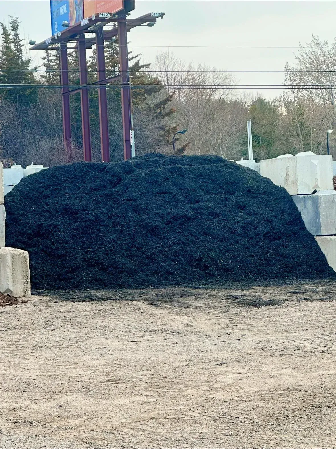 Large pile of black mulch at a garden supply store, in front of concrete blocks and a billboard.