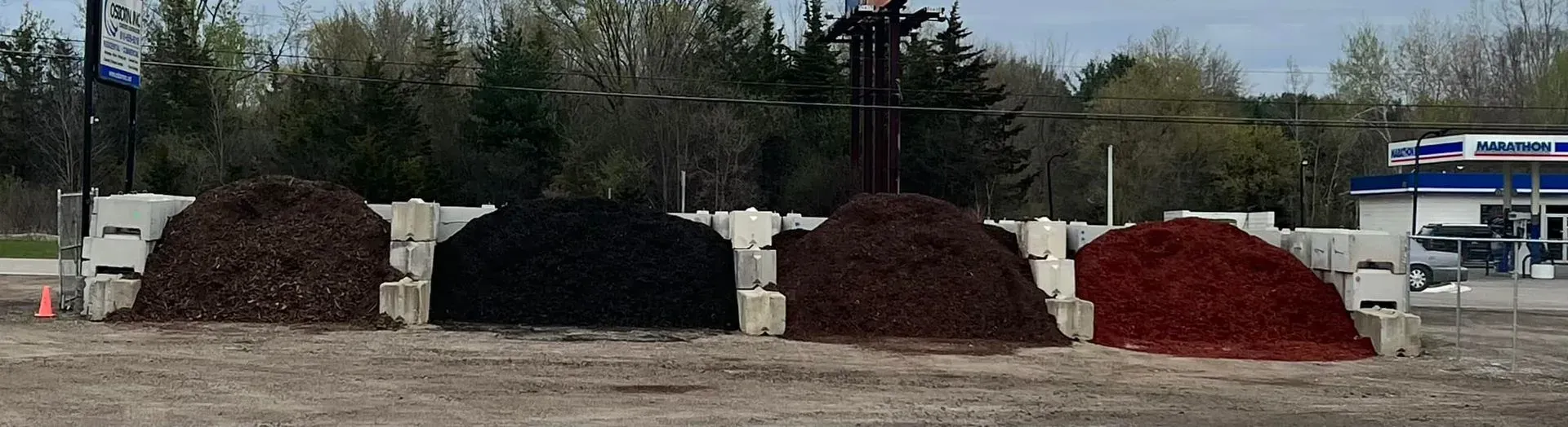 Piles of different colored mulch sit in front of concrete barriers, trees in the background.