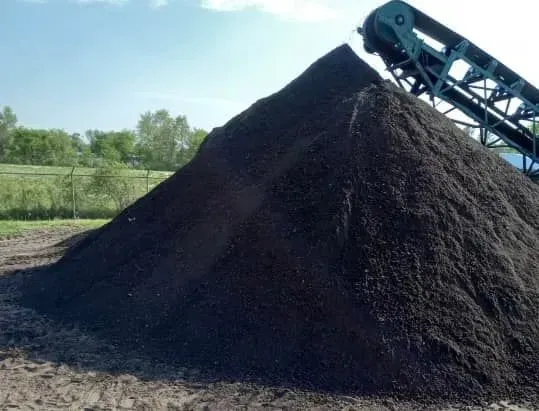 Large pile of dark compost material next to a conveyor belt; outdoors on a sunny day.