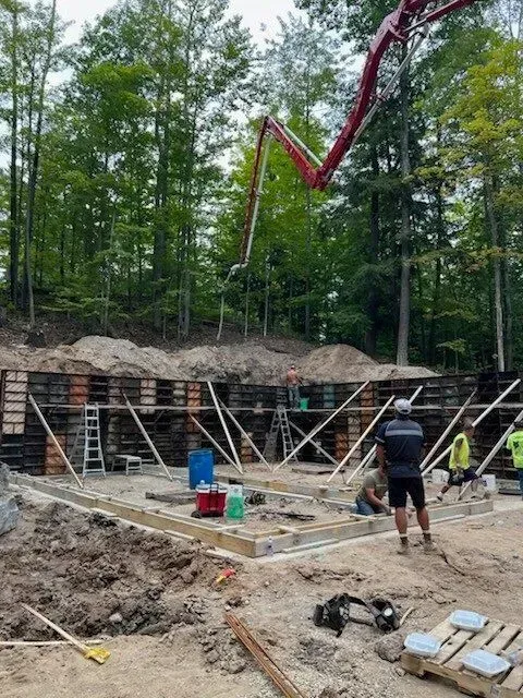 Construction site with workers, concrete forms, and pump truck, set against a wooded hillside.