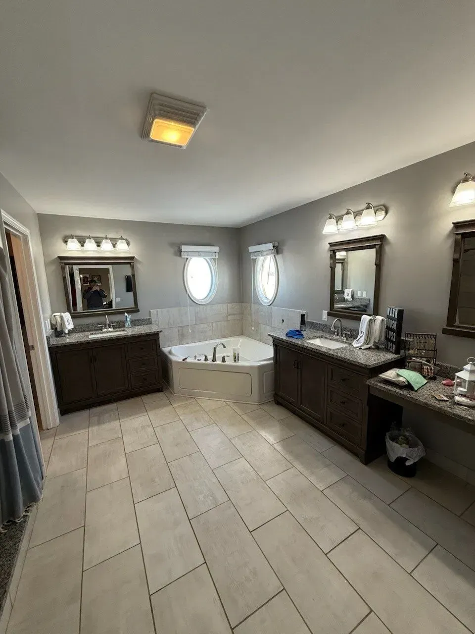 Spacious bathroom with beige tile floors, dark wood vanities, a jacuzzi tub, and two oval windows.