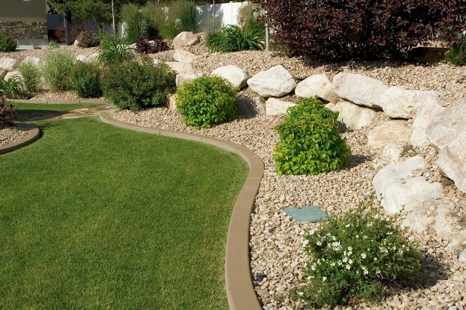 Lush green lawn with curving border, surrounded by gravel, rocks, and various shrubs and plants.