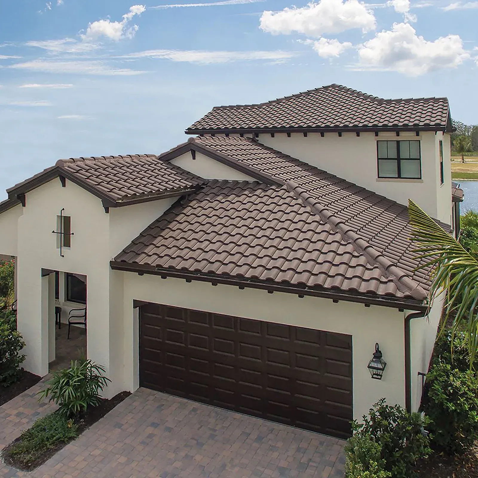 Two-story stucco house with brown tile roof and garage. Sunny day with landscaping and water view.