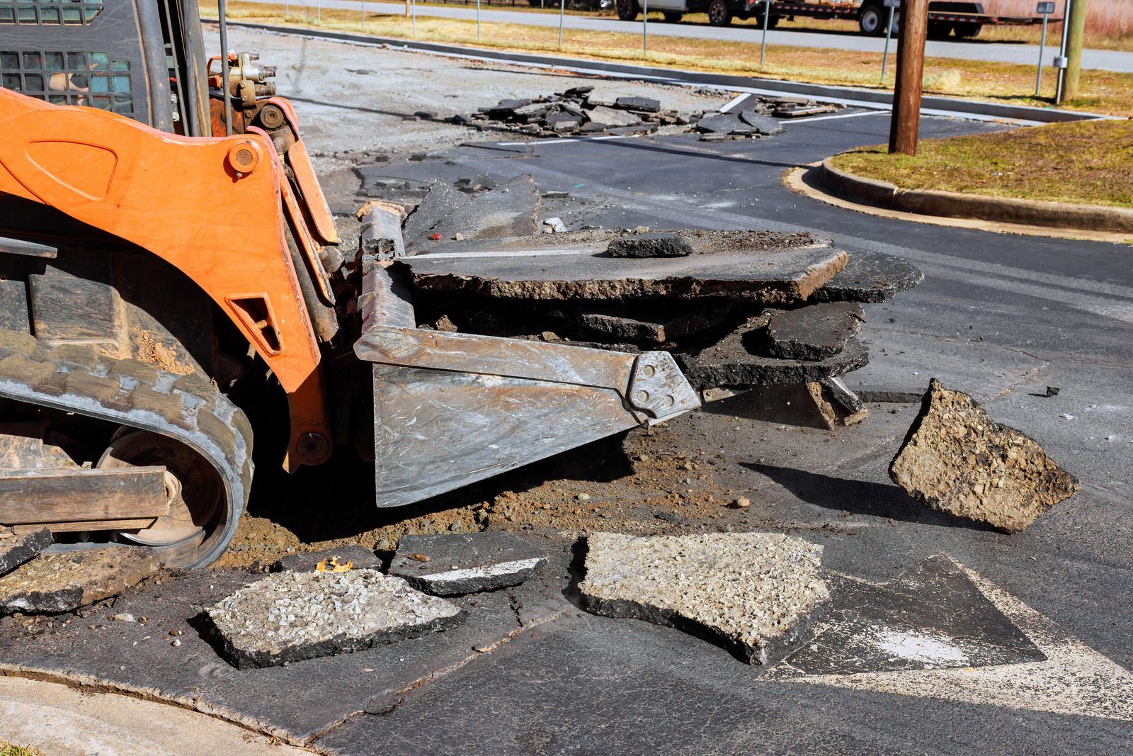 Orange skid steer removing broken asphalt from a street, debris scattered.