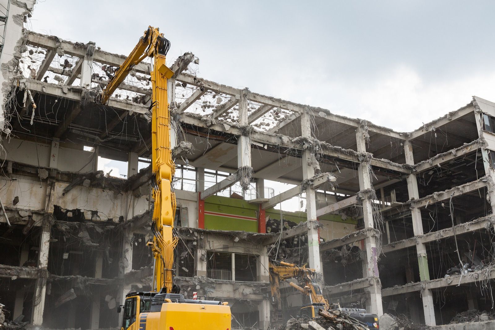 A yellow excavator demolishes a multi-story building. Debris and dust fill the air.