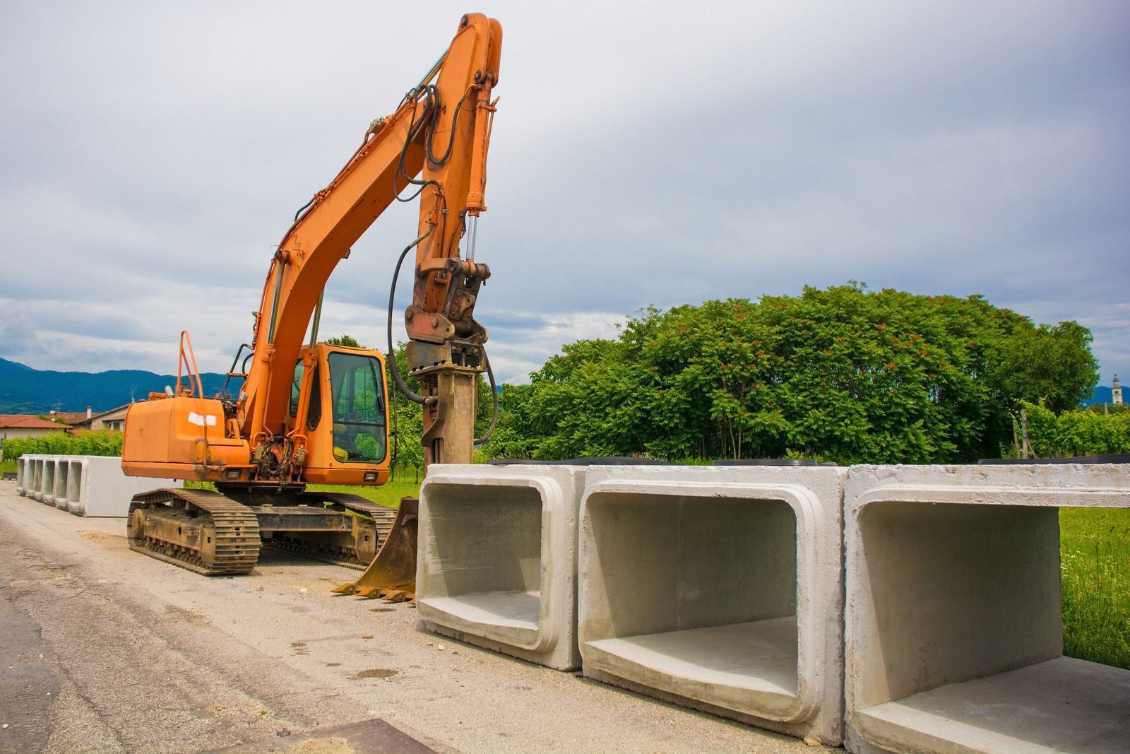 Orange excavator next to concrete culverts on a road, with greenery and cloudy sky in the background.