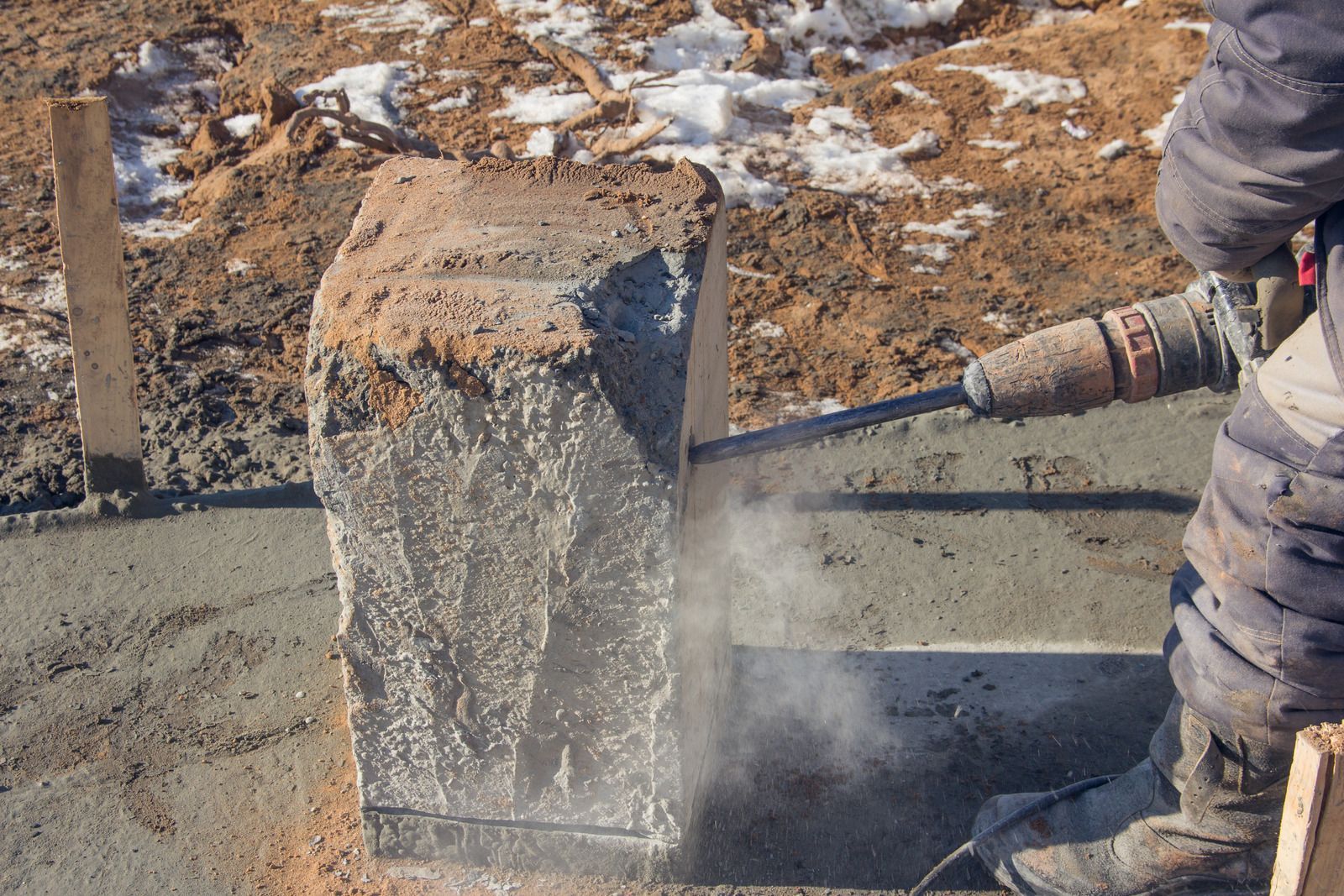 Construction worker using a jackhammer to cut a large concrete block in a construction site, releasing dust.