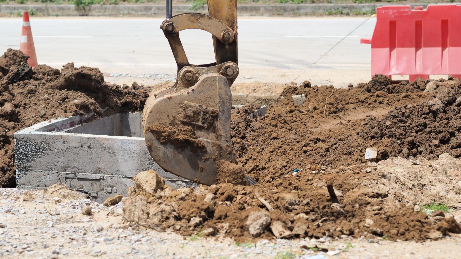 Excavator digging near a concrete structure on a construction site; dirt pile on the ground.