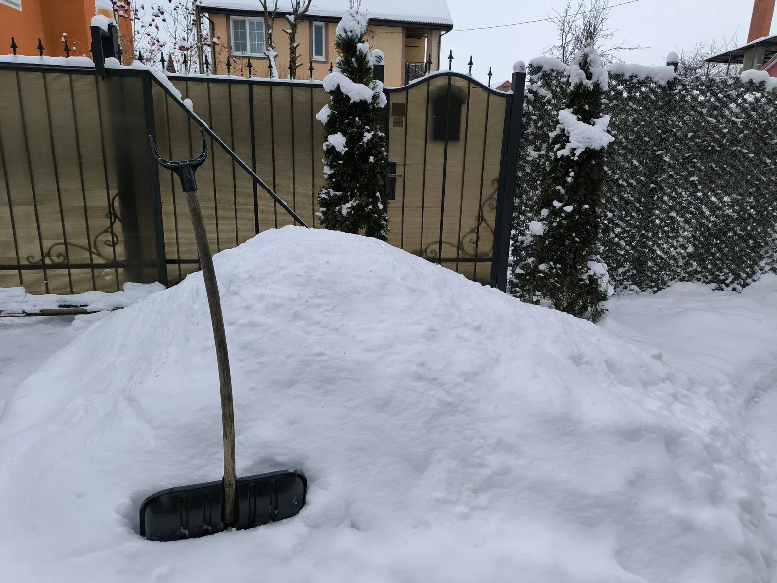 Snow pile with shovel leans against a black fence in a snowy outdoor setting.