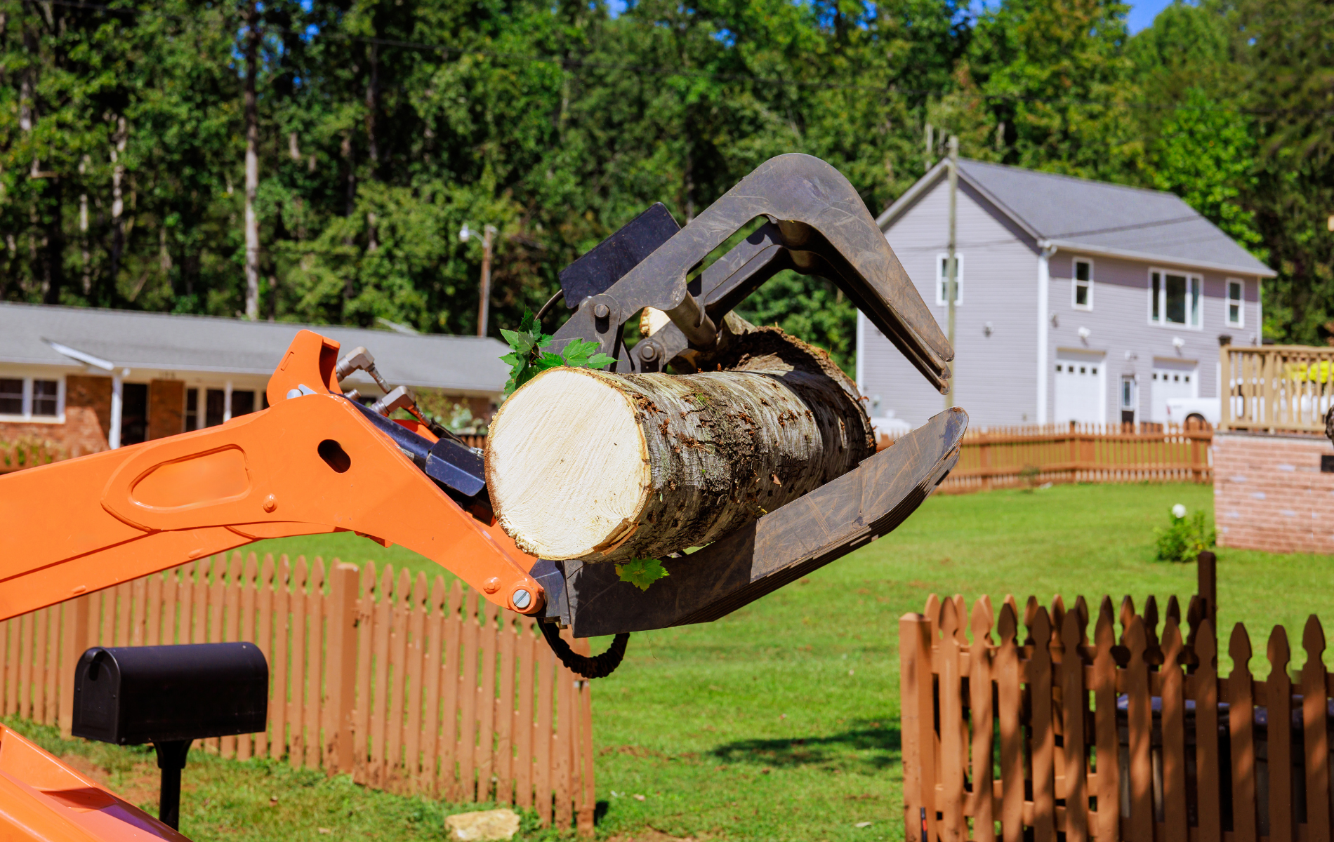 Orange machinery lifting a tree log in a residential yard. A house and wooden fence are in the background.