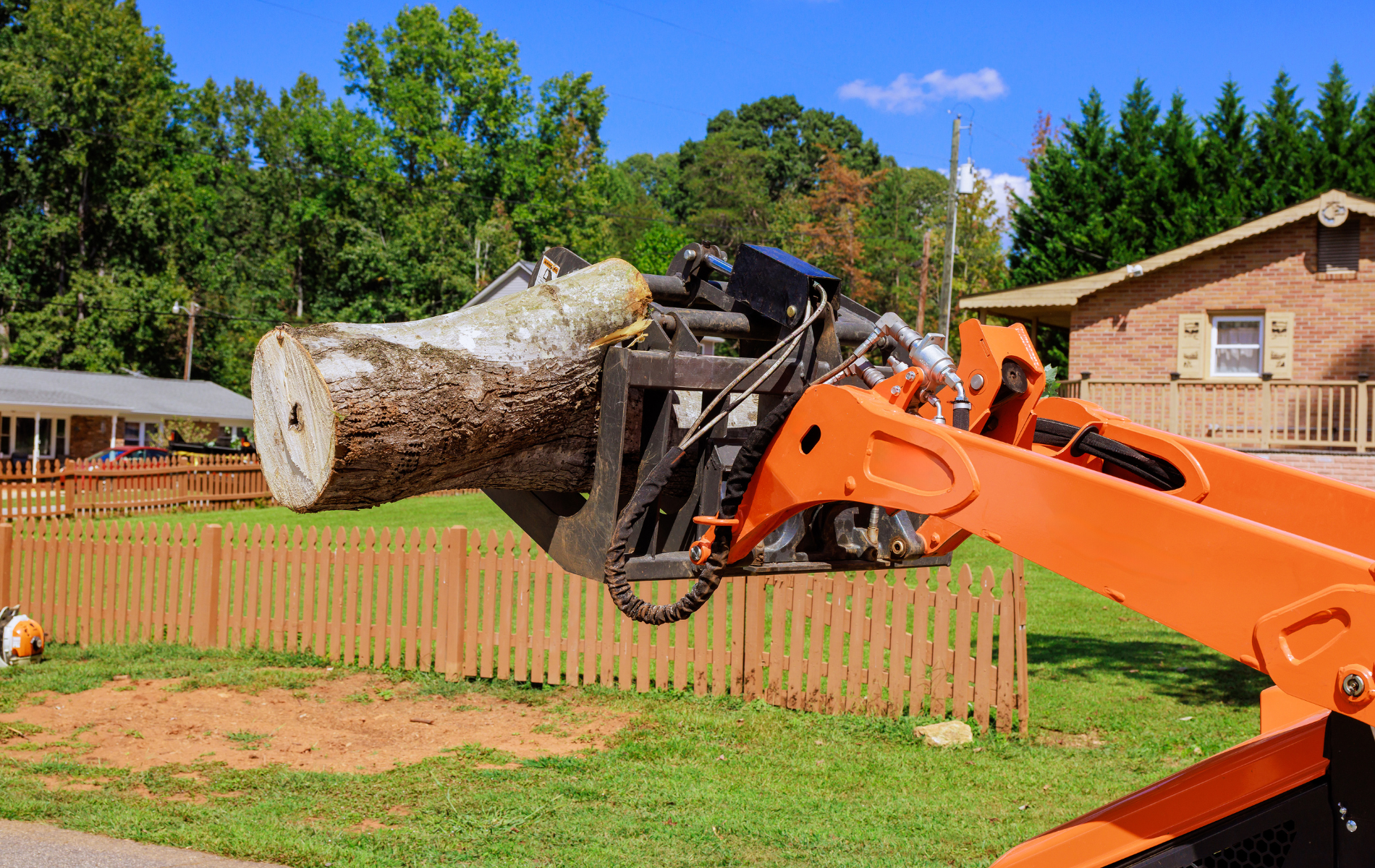 Orange loader carrying a large tree trunk over a wooden fence, near a house and trees.