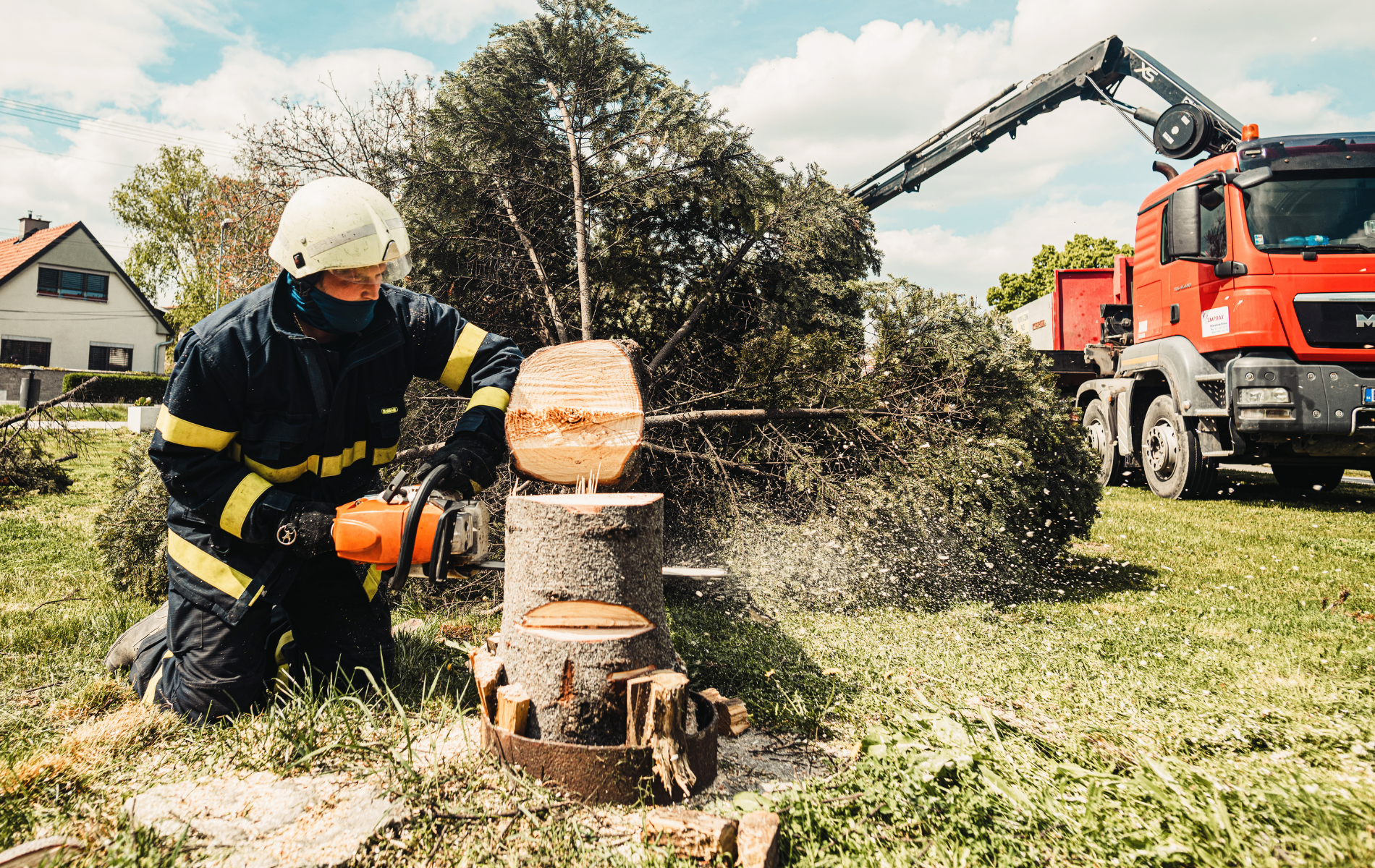Firefighter using a chainsaw to cut a tree trunk; a crane truck is in the background.