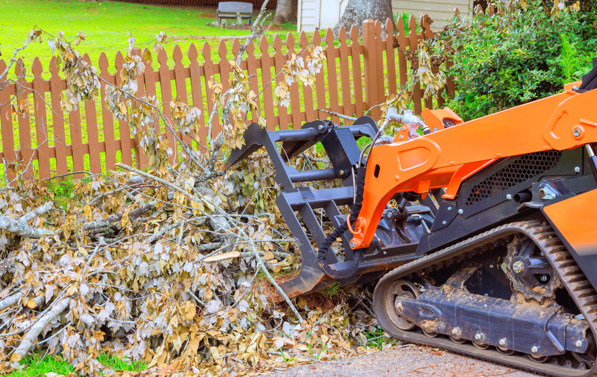 Orange track loader moving brush next to a brown wooden fence.