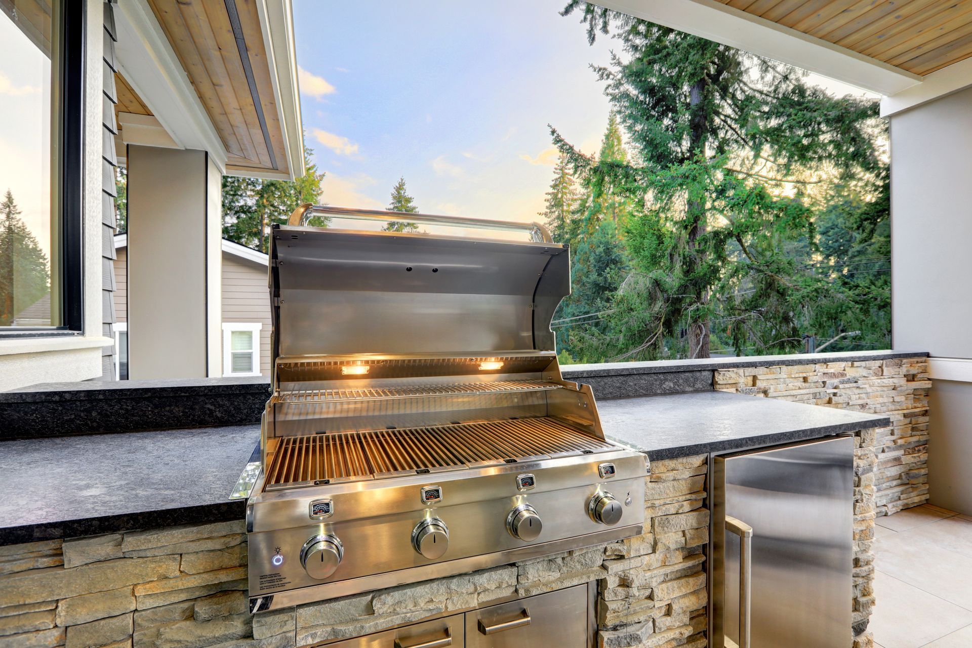 Stainless steel outdoor grill integrated into a stone countertop on a covered patio overlooking trees.