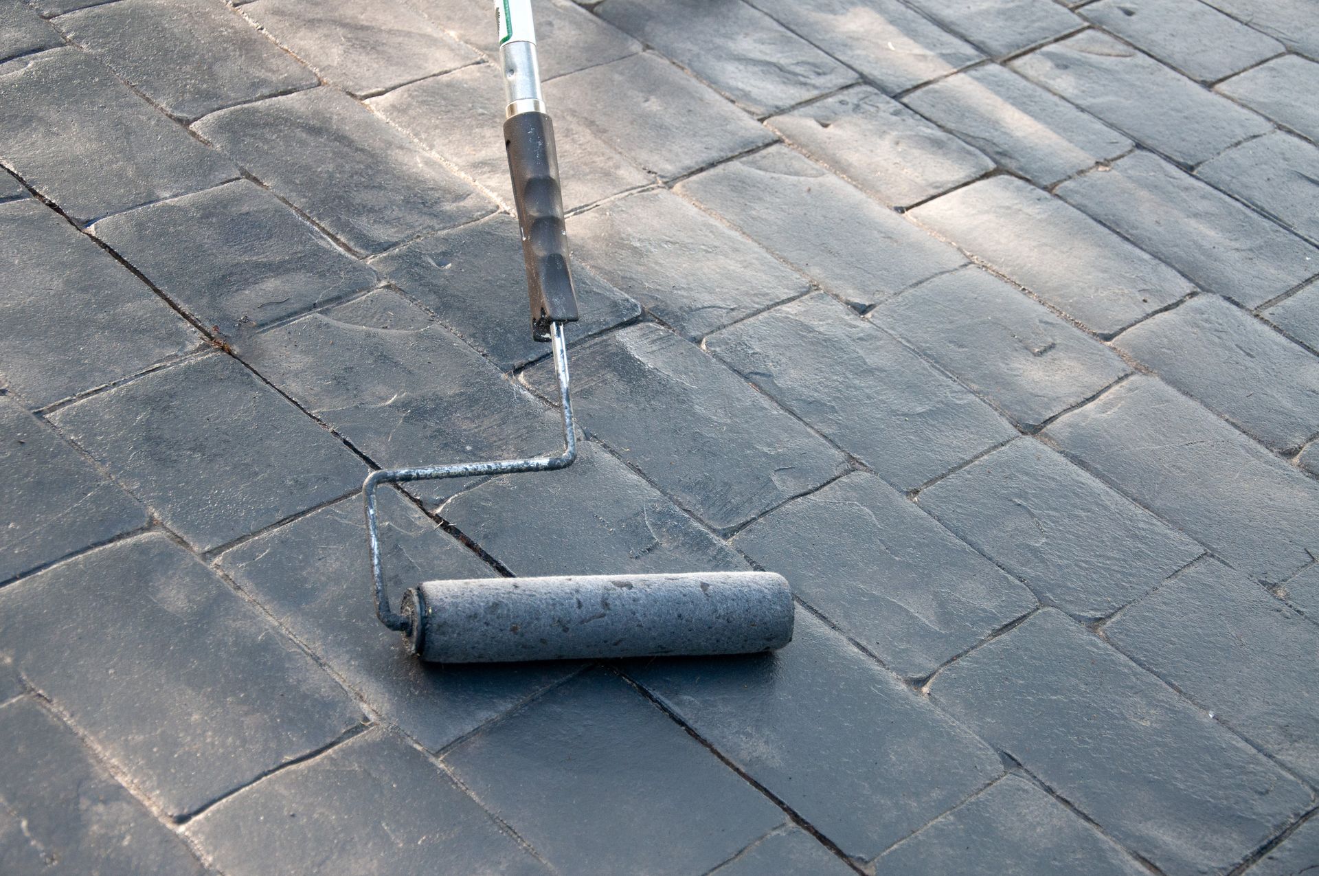 A paint roller applying sealant to dark gray concrete pavers.