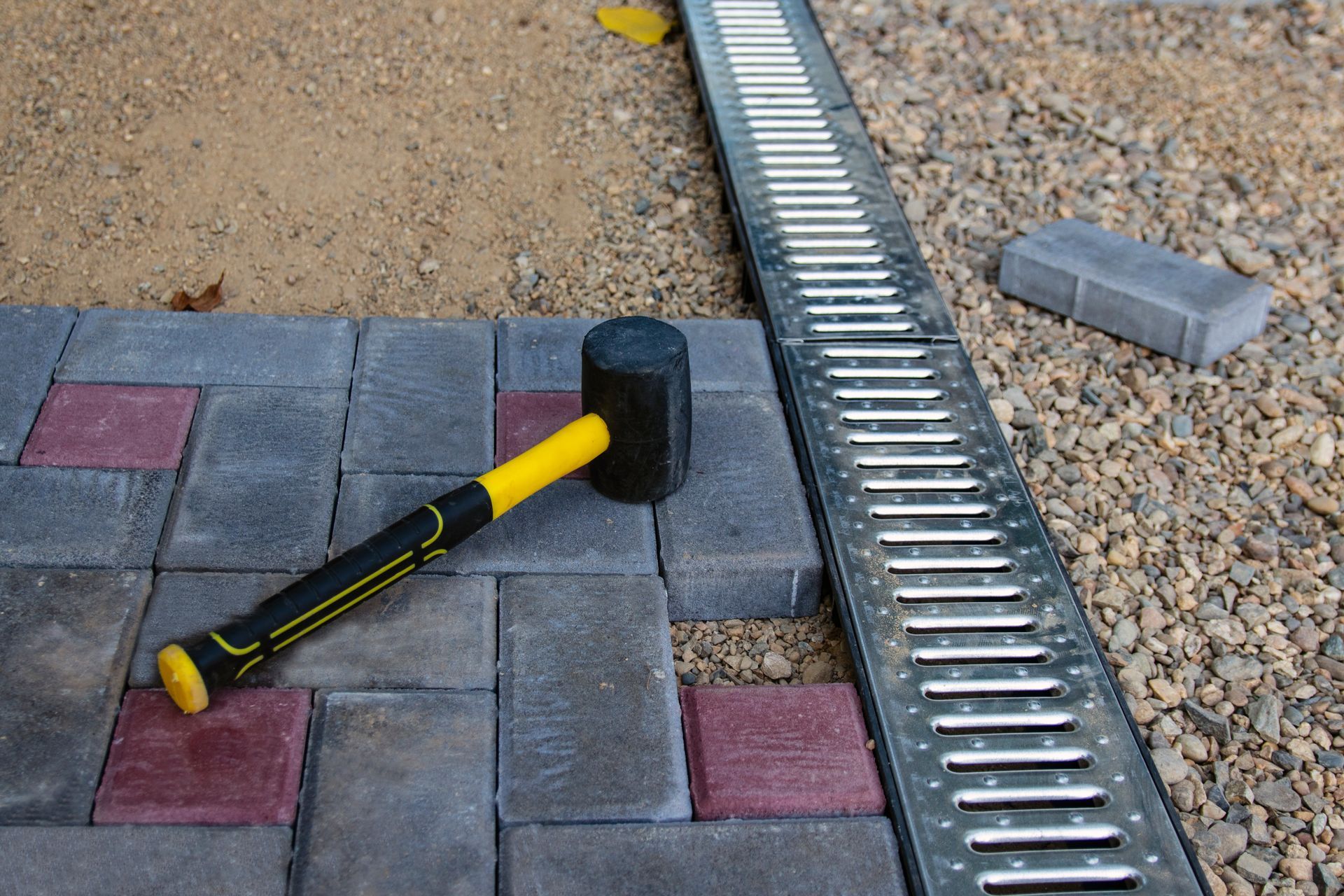 A rubber mallet lies on top of gray and red paving stones next to a metal drainage grate on a gravel path.