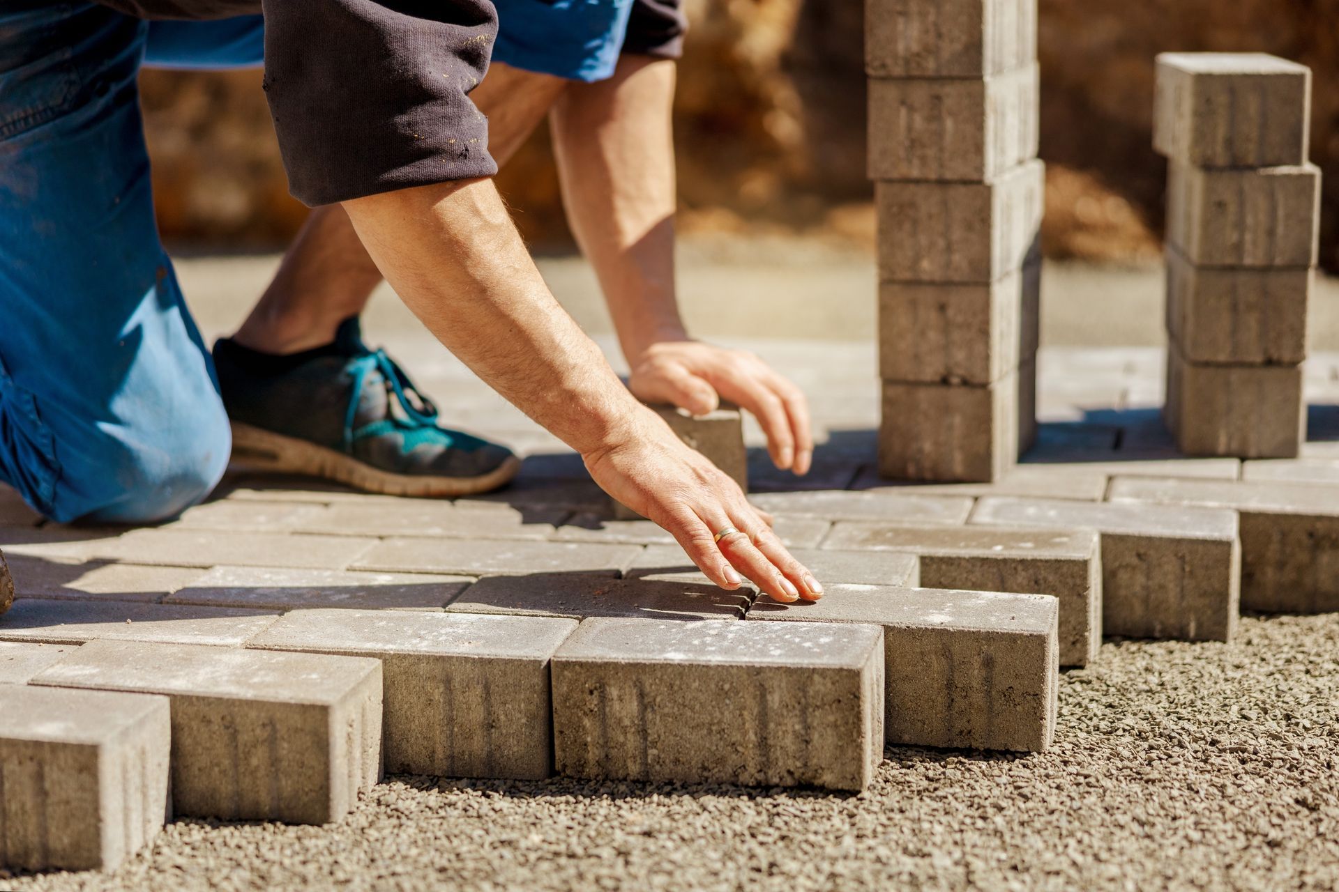 A person in work clothes kneeling on the ground while placing rectangular paving bricks into a pattern.