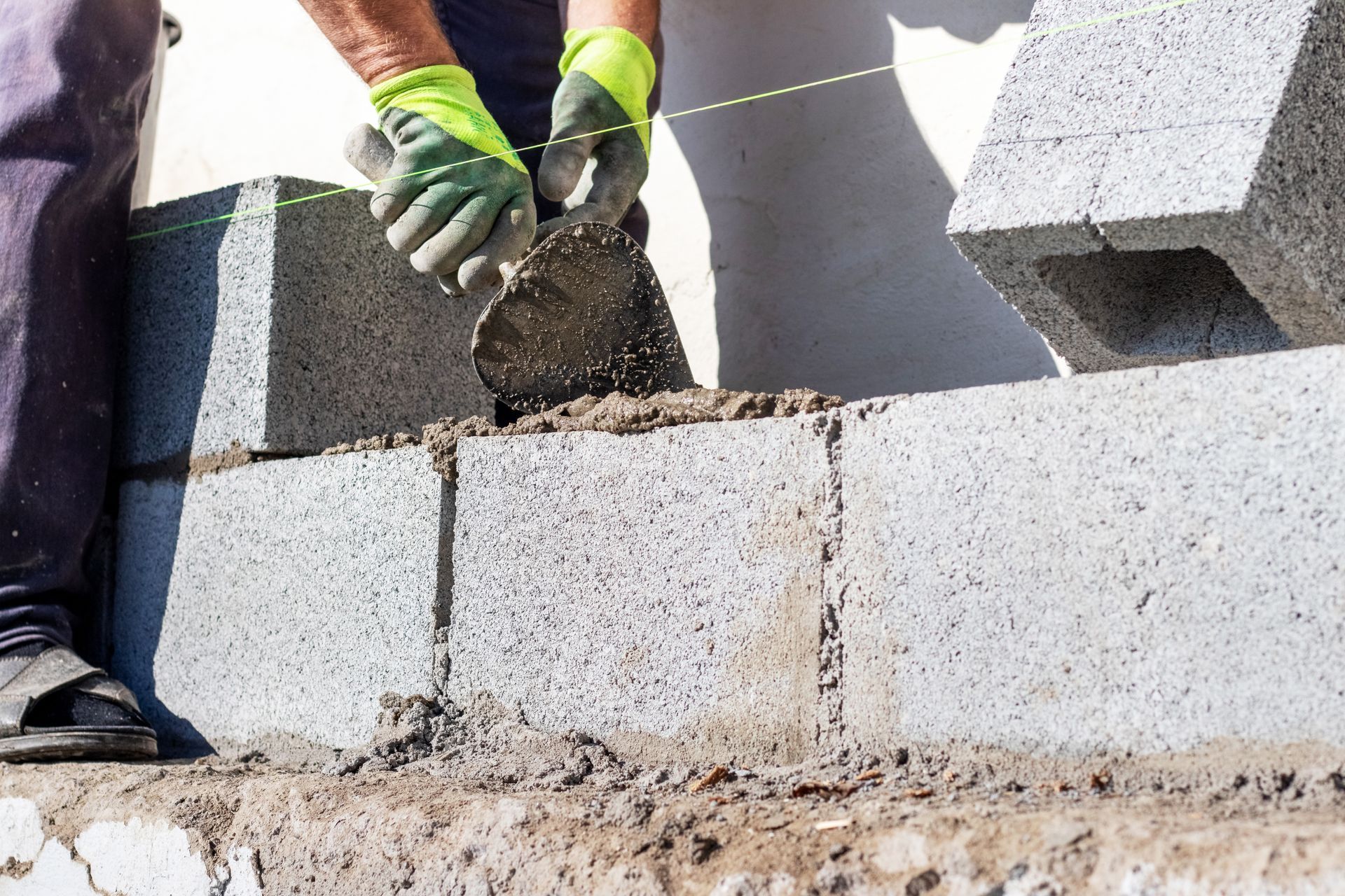 A worker in lime green gloves uses a trowel to apply mortar to gray concrete blocks being laid in a wall.
