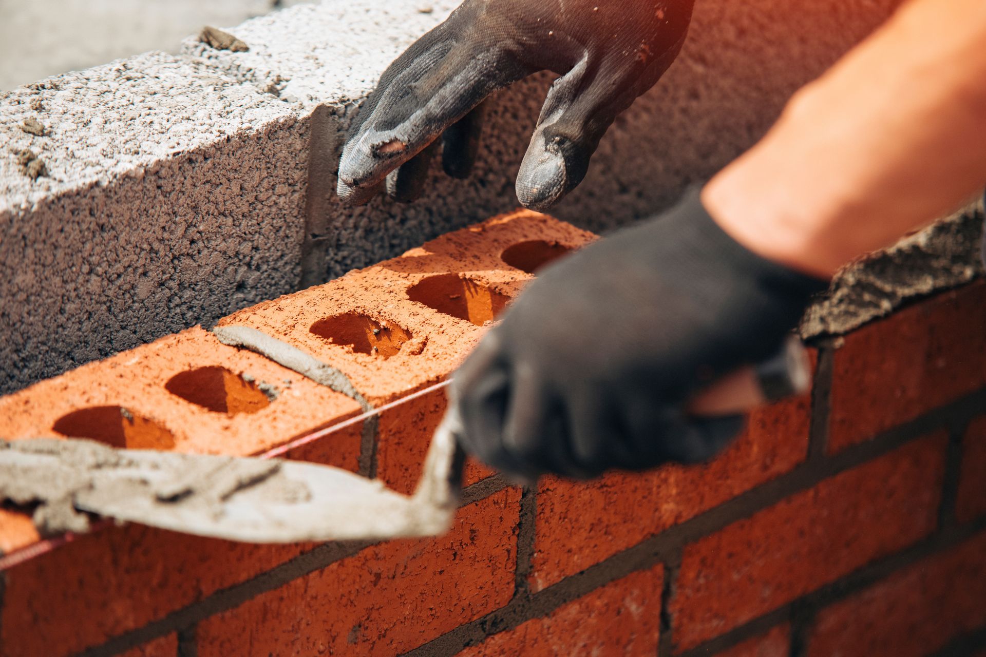 A gloved hand holds a trowel to apply mortar to a row of orange bricks while another hand positions a new brick.