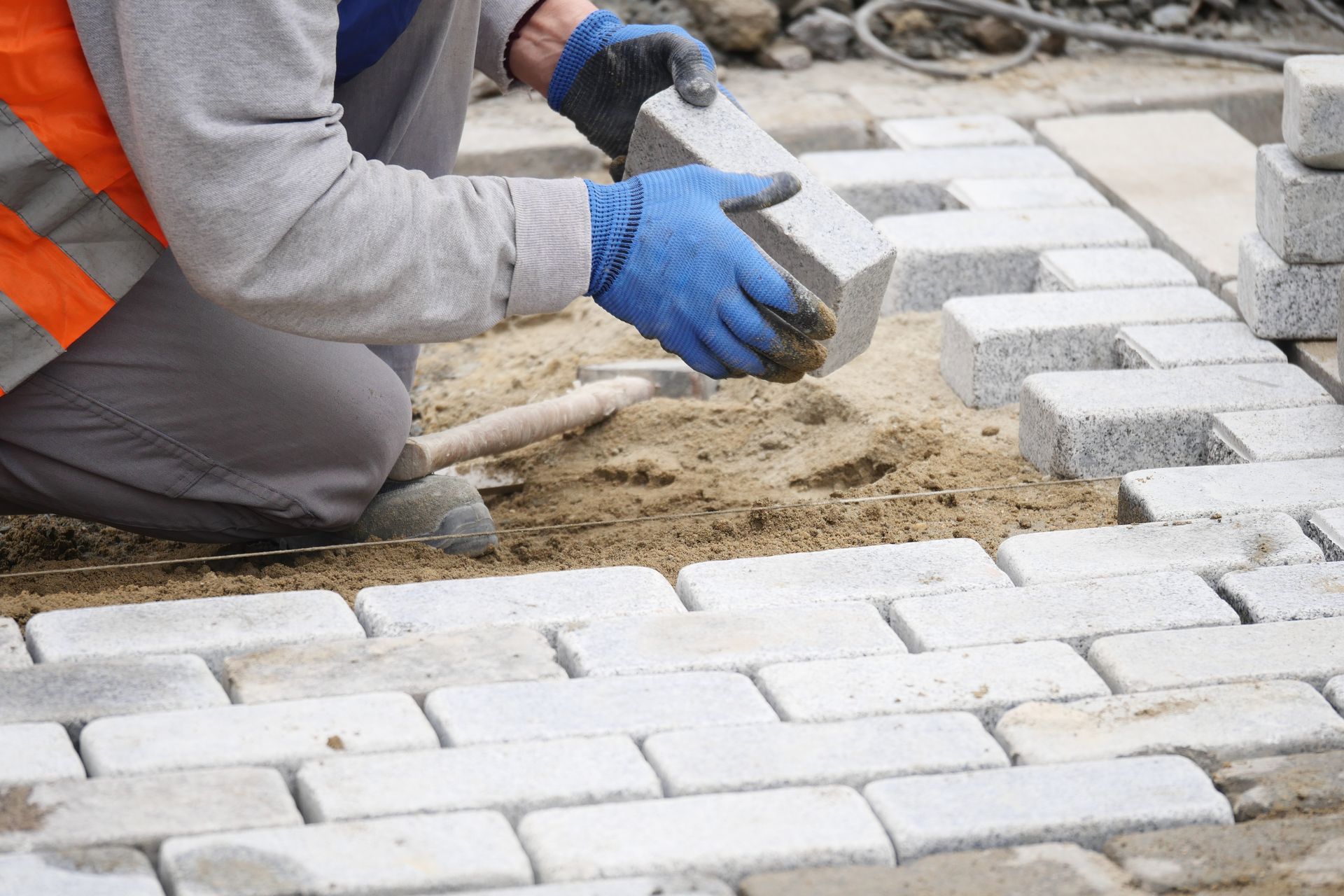 A construction worker in a high-visibility vest and blue gloves places a stone paver into a sand bed.