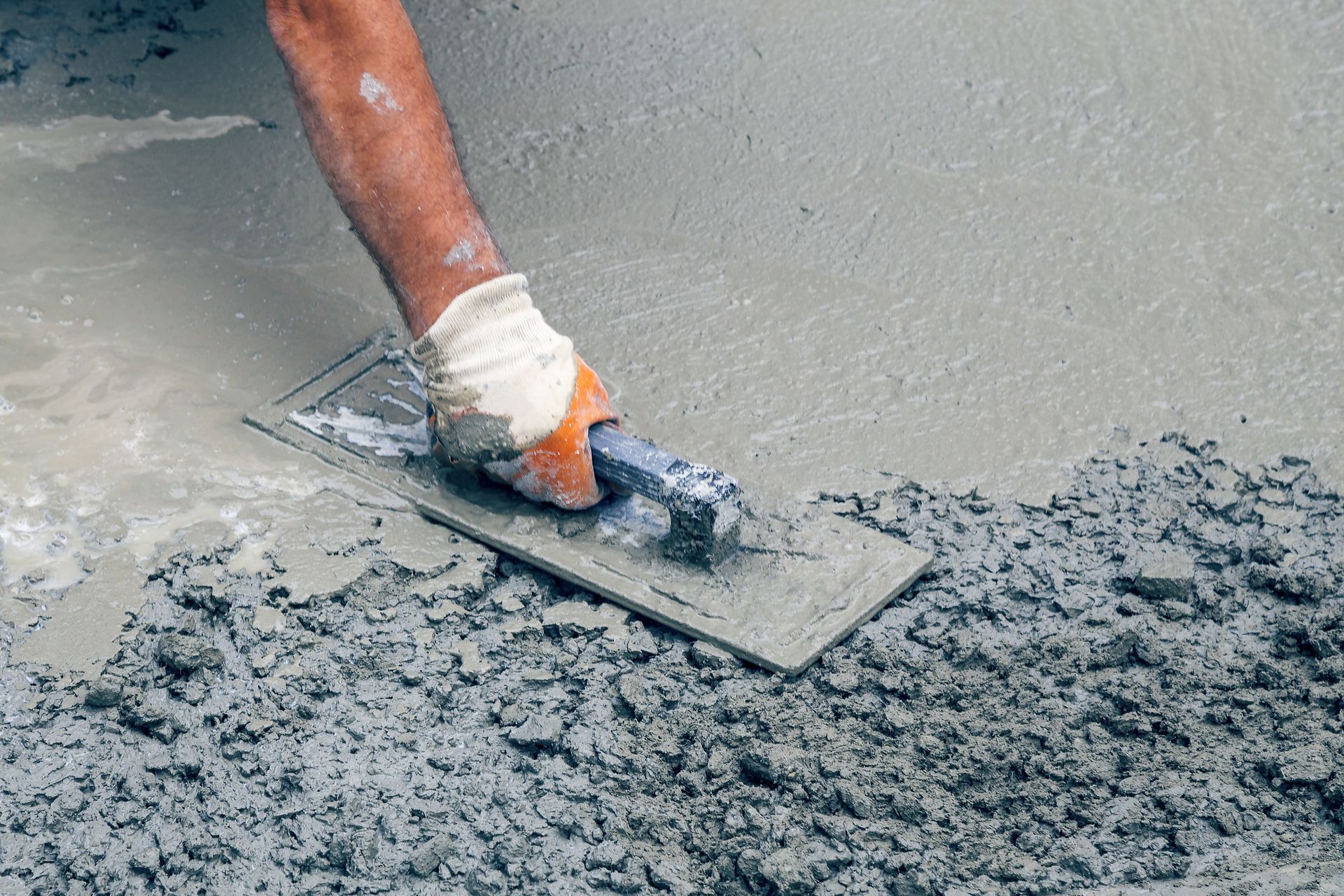 A person wearing a glove uses a hand float to smooth wet concrete on a construction site.