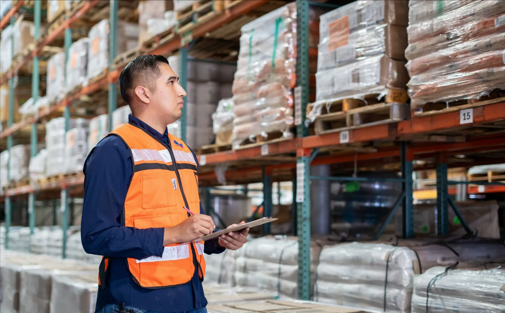 Supervisor con chaleco de seguridad naranja realizando conteo de inventario con tablilla en almacén industrial con estanterías de paletas numeradas