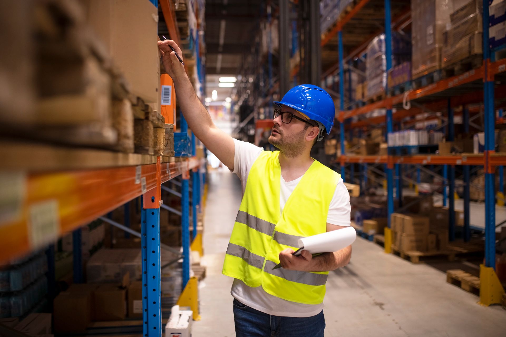 Warehouse worker in blue hard hat and yellow vest checking items on a shelf in a warehouse
