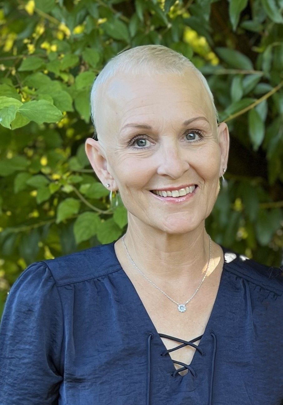 A woman with a shaved head is smiling in front of a tree.