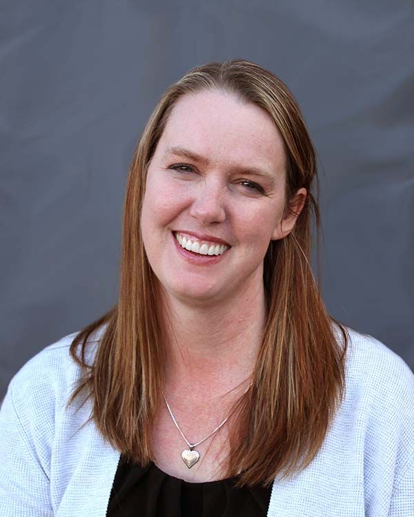 A woman wearing a white sweater and a heart necklace is smiling.