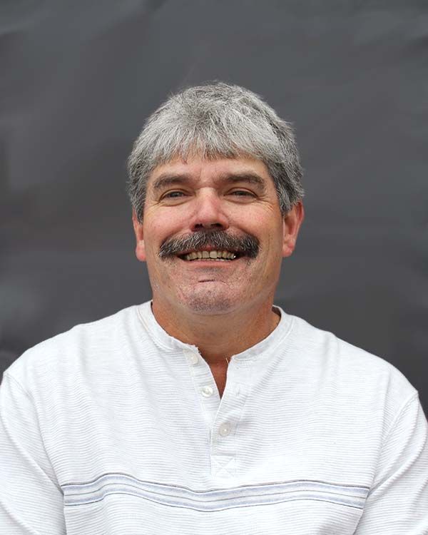 A man with gray hair and a mustache is smiling and wearing a white shirt.