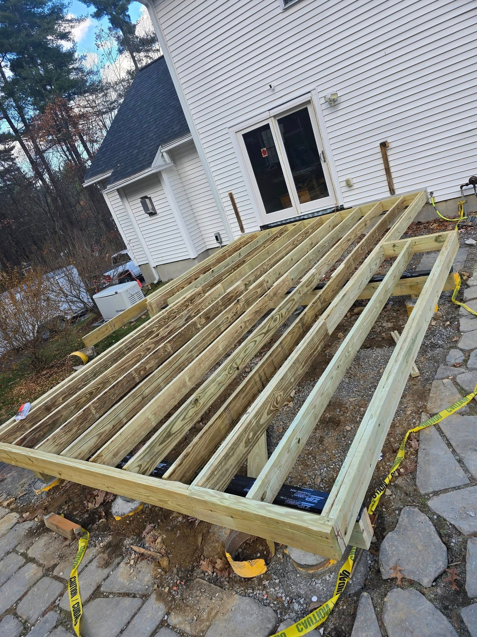 Wooden deck frame under construction near a white house with stone pavers. Yellow caution tape surrounds the site.