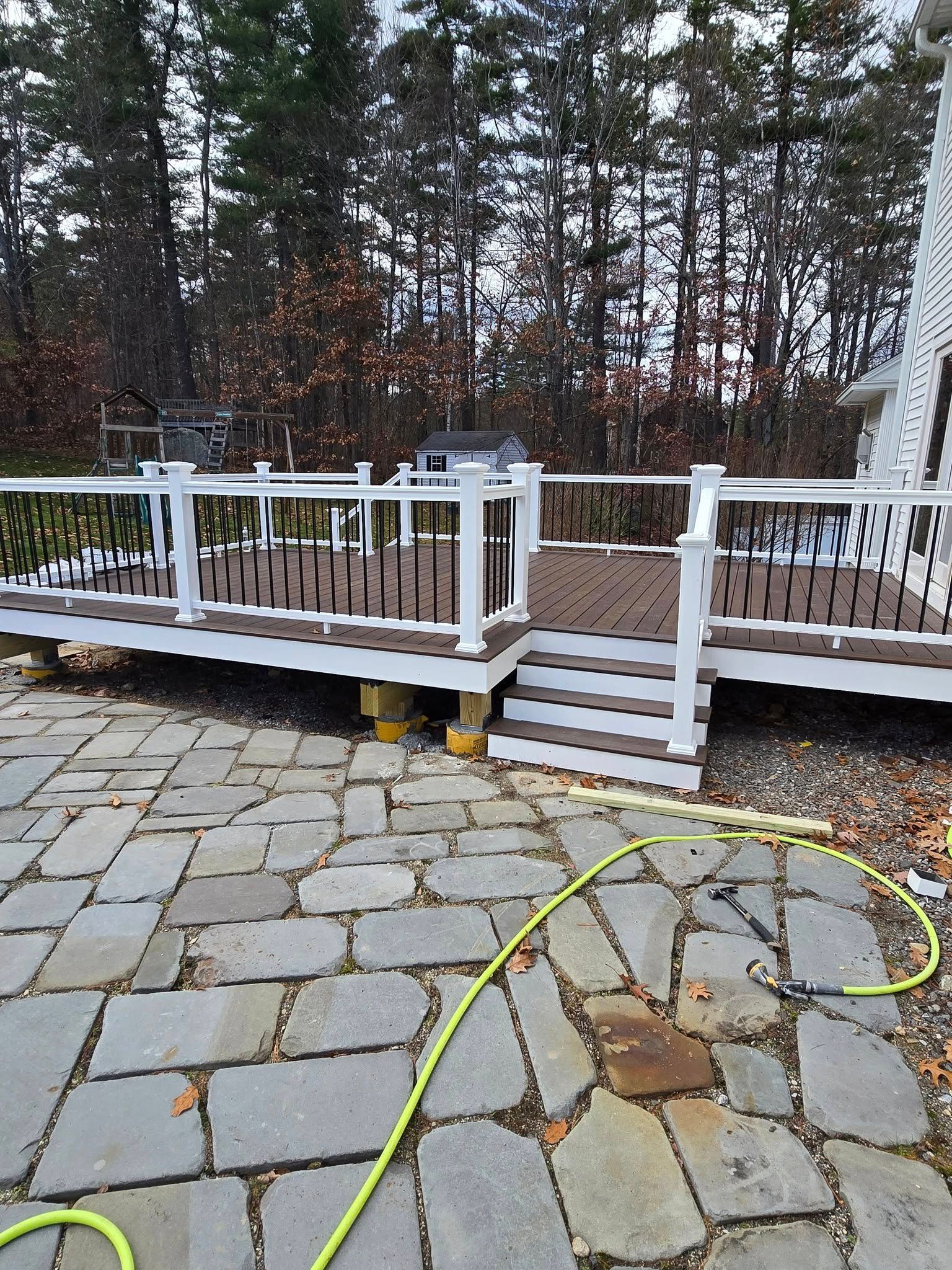 Outdoor deck with white railing and brown decking, over a stone patio. Hose and trees visible in background.